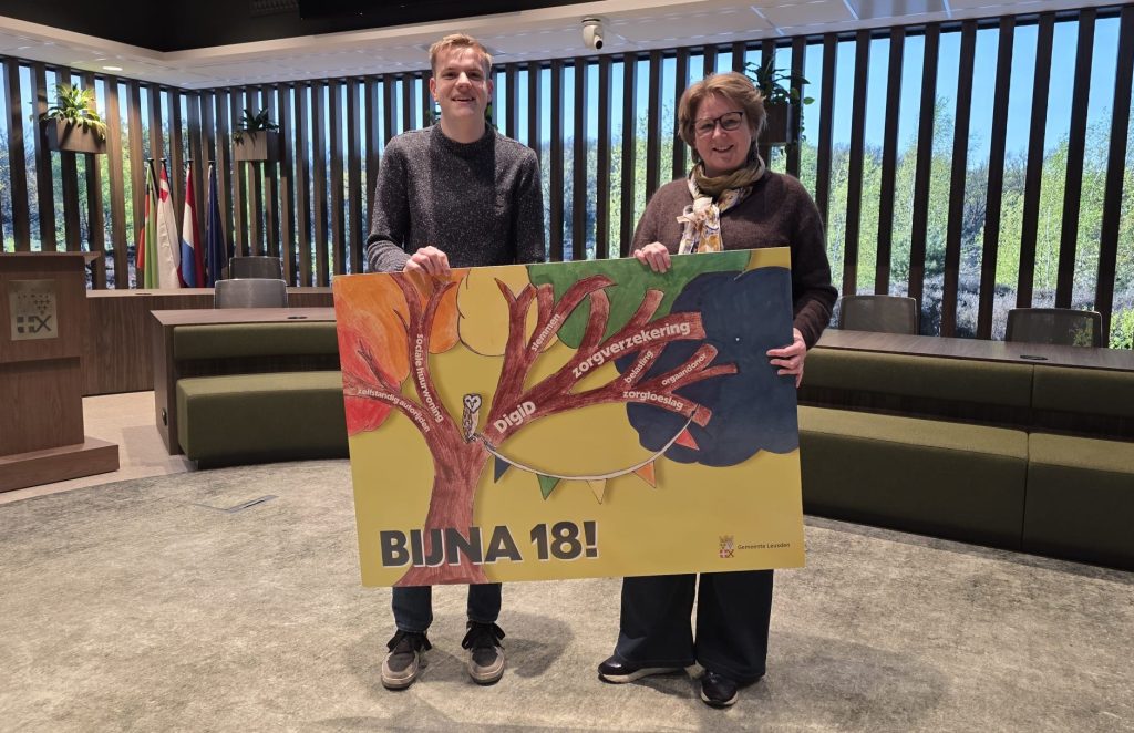 Thomas en wethouder Marleen Treep staan in de raadzaal van het gemeentehuis en houden samen een groot exemplaar van de verjaardagskaart voor 18-jarigen vast. Op de kaart zie je een boom met takken, waarop woorden staan zoals 'zorgverzekering' en 'DigiD' en vetgedrukt de tekst "Bijna 18!".